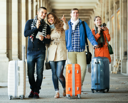 Two couples of young smiling travelers with luggage and cameras walking by street in the city. Selective focusの写真素材