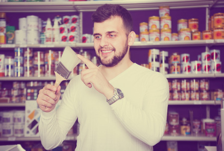 cheerful male customer examining various types of brushes in paint supplies storeの写真素材
