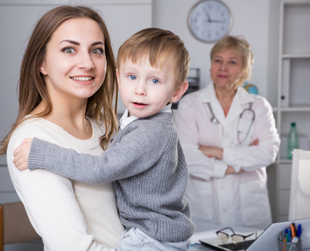 Smiling female is standing satisfied with her toddler after visiting clinic indoor.の写真素材