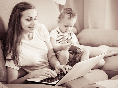 Satisfied mom is productively working behind laptop while kid playing on tablet at home.の写真素材