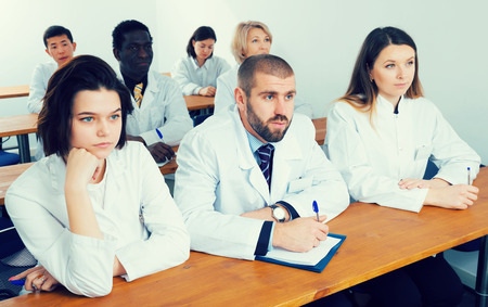  positive group of people in white coats attentively listening and making notes while sitting in boardroomの写真素材