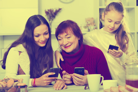 Modern grandmother with adult daughter and granddaughter, sitting with smartphones in home interiorの写真素材