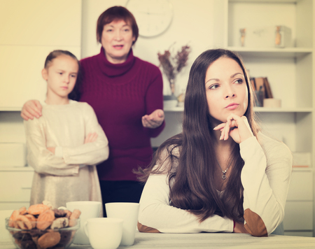 Portrait of young mother upset after quarrel with little daughter standing behind with grandmaの写真素材