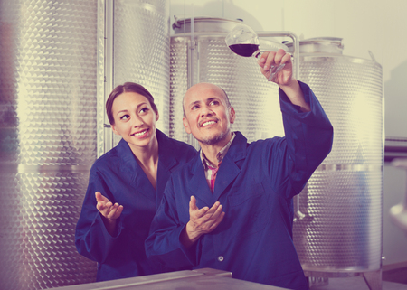 Happy positive two factory workers in uniform examining sample of wine in glassの写真素材