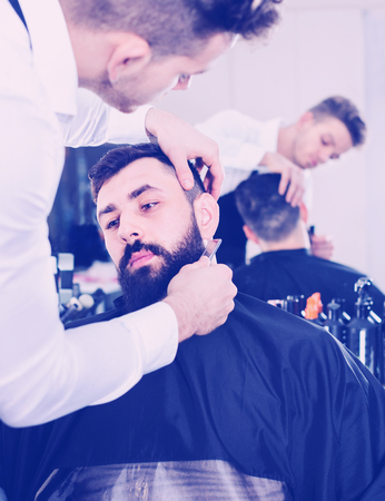 Pleasant  male hairdresser accurately cutting beard of client at hair salonの写真素材