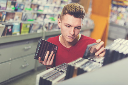 Closeup serious young man absorbedly choosing CD and DVD in shopの写真素材