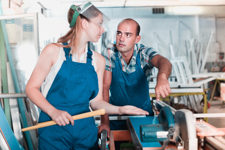 Experienced worker helping diligent friendly smiling girl in work on circular saw at plastic windows factoryの写真素材