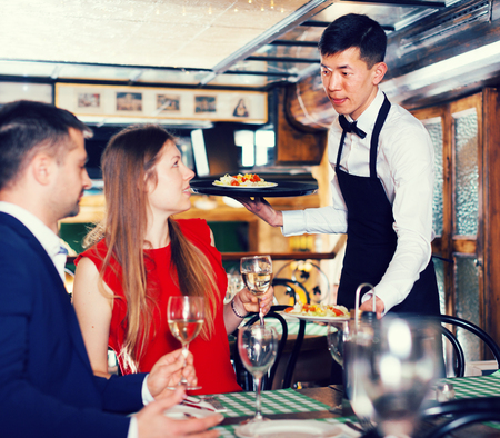 Male waiter is serves an order for smiling couple who is drinking wine and eating in restaurant.の写真素材
