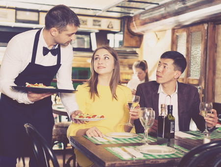 Male waiter is serves an order for smiling couple who is drinking wine and eating in restaurant.の写真素材