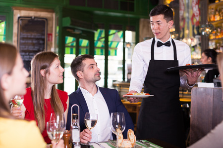 Man waiter is brings order to friends who have dinner and drink wine in restaurant.の写真素材