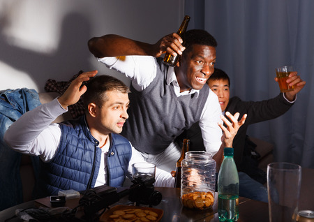 Exalted male friends watching tv together at home, enjoying beer with snacks indoors late at nightの写真素材