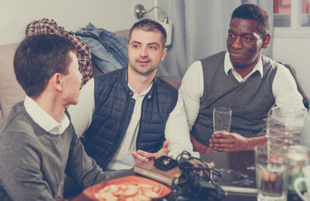 Three cheerful male friends meeting and discussion with beer and snacks at home