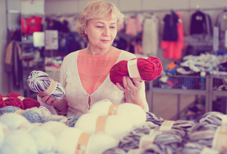 Positive smiling mature woman buyer choosing colored yarn for knitting on saleの写真素材