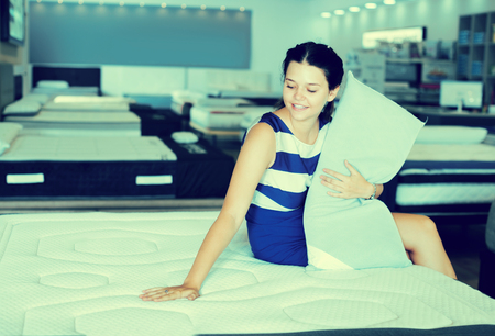 Smiling girl sitting with pillow at new mattress in furniture storeの写真素材