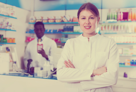 Experienced cheerful positive smiling female pharmacist standing on background with male colleague in pharmacyの写真素材