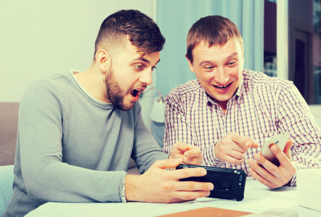 Happy male friends using phone conversing with liveliness at home table