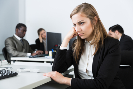 Upset thoughtful girl sitting at workplace in modern coworking space on background with working colleaguesの写真素材