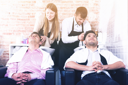 Men leaning heads on sink and relaxing while hairdressers washing their hair in barbershopの写真素材