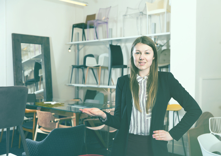 Confident woman seller of furniture shop inviting customers to chairs departmentの写真素材