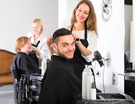 Handsome man sitting in a barbershop wearing a peignoir is waiting for beautiful hairdresser to make him a stylish haircutの写真素材