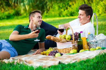 Cheerful young guy with friend sitting on green lawn drinking wine on picnicの写真素材