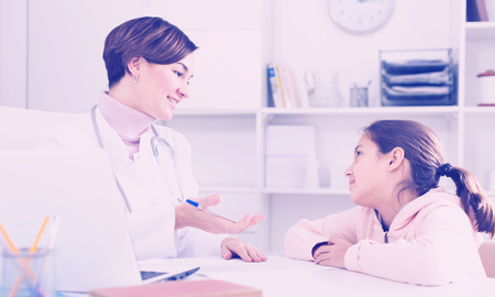 Doctor fills patient's card for girl and asks her about health in medical officeの写真素材