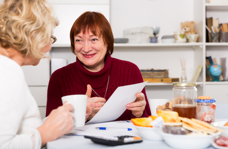 Two positive mature women considering papers while sitting on kitchenの写真素材