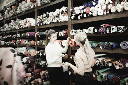 Polite female seller helping woman in choice of interesting cloth in textile shopの写真素材