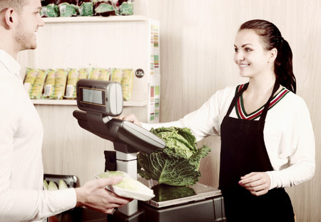 Smiling woman seller helping customer to weigh cabbage in grocery shop