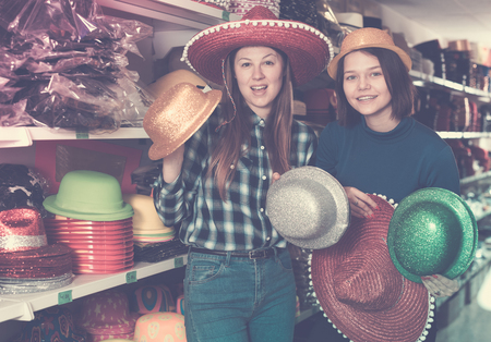 Two cheerful young female friends preparing for party, choosing funny headdresses in storeの写真素材