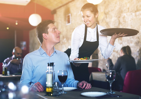 Positive waitress bringing delicious meal to young male in cafeの写真素材