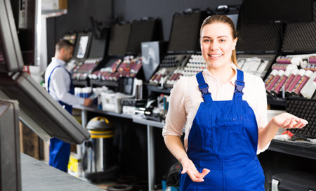 Positive girl in workwear standing near stand with paints in car repair shop, meeting clients