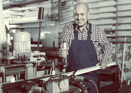 Positive smiling middle-aged man in coverall toiling on a machine in PVC shop