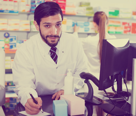 Positive man pharmacist is writing to note inventsry of medicines near computer in pharmacyの写真素材