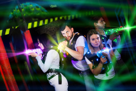 Modern young people playing laser tag on dark labyrinth in bright beams of laser pistols