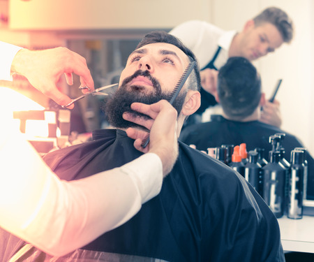 Glad stylist creating new shape for beard of client at hairdressing salonの写真素材