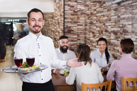 Cheerful male waiter serving rural restaurant guests at a tableの写真素材