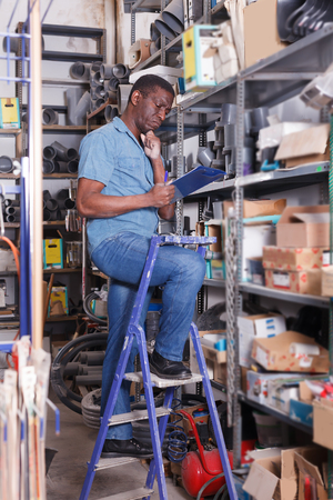 Nice African American male warehouse worker making notes while performing inventory of building materialsの写真素材