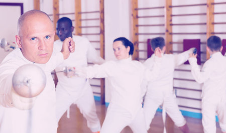 Portrait of adult focused man wearing fencing uniform practicing with foil in gym
