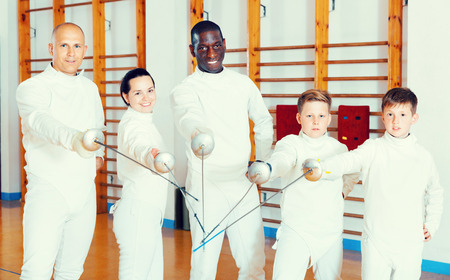Group portrait of young cheerful positive fencers with coaches holding rapiers in training roomの写真素材