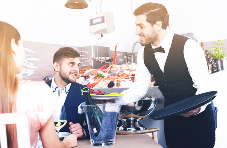 smiling waiter bringing ordered seafood dishes to smiling couple at fish restaurantの写真素材