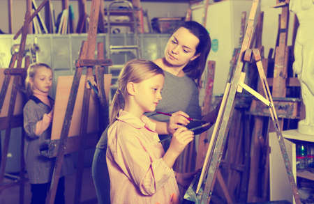 Skillful woman teacher correcting work of girl during painting class at art studioの写真素材