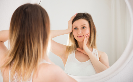 Young woman using mirror and touching face in bedroom in morningの写真素材