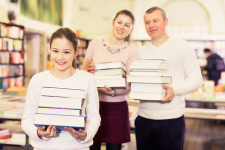 Positive smiling preteen girl and her parents holding piles of books bought in bookstoreの写真素材