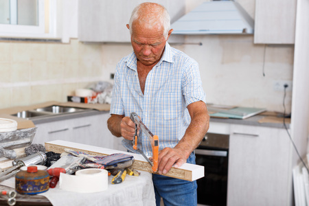 Skillful older man engaged on his home renovations, cutting wood chipboard plank with hand sawの写真素材