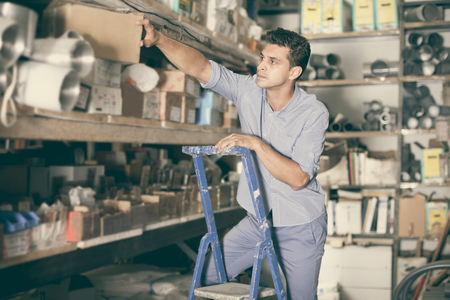 Positive male in unifom is standing on ladder near shelves in the building storeの写真素材