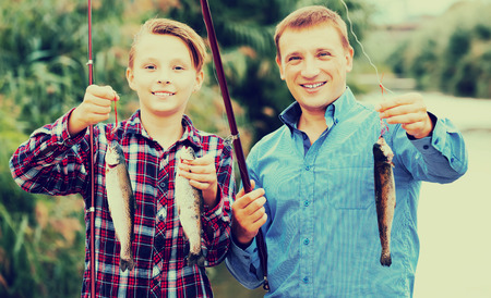 Cheerful teenage boy and his father holding fish on hook in handsの写真素材