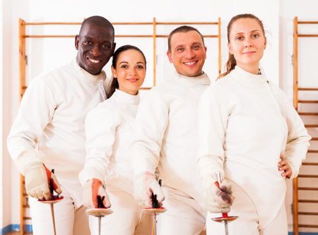 portrait of happy men and women fencers posing with foils  together at fencing room