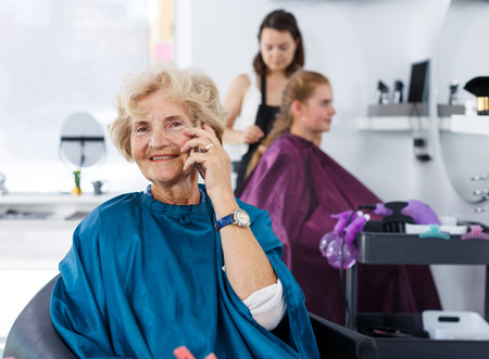 Portrait of elderly female client waiting for hair styling in modern hair salon, talking on phoneの写真素材