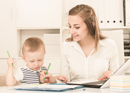 Woman 22-27 years old is working on laptop while child painting on papers in office.の写真素材